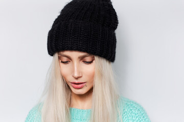 Close-up studio portrait of young pretty girl with blonde hair wearing black hat and blue sweater on white background.