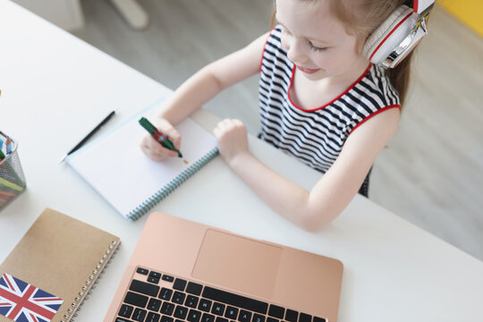 Little girl in headphones sitting in front of laptop and writing in notebook
