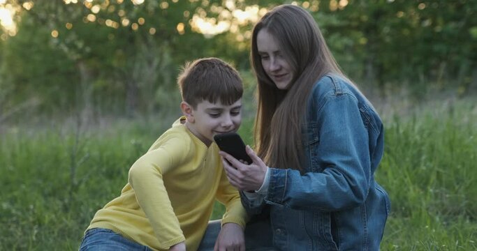 Young mother shows photo on phone to her son. Mom and son in park at picnic.