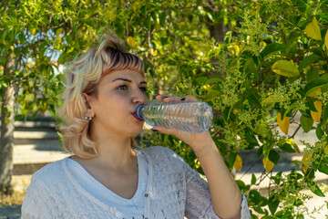 chica con blusa blanca bebiendo agua de una botella de plástico, chica de pelo corto y flequillo bebiendo agua en el parque