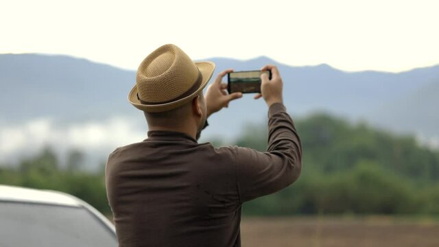 A young Asian man drives a beautiful nature drive. He was standing in front of the car on the roadside. He uses his phone to take pictures of beautiful views.