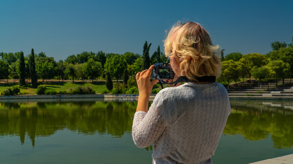 Chica rubia de espaldas haciendo fotos con cámara réflex a lado del lago, mujer fotógrafa haciendo fotos en el parque