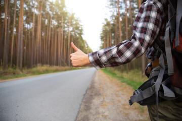 Hand with thumbs up on the background of highway - hitchhiking, voting. Tourist in a check shirt with backpack near the forest hitch a ride. Backpacker, domestic tourism, adventure alone, trip, hike