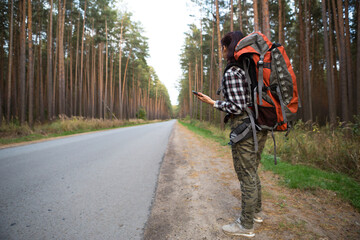 Female tourist in a check shirt with an orange large backpack near a highway in the woods with a...