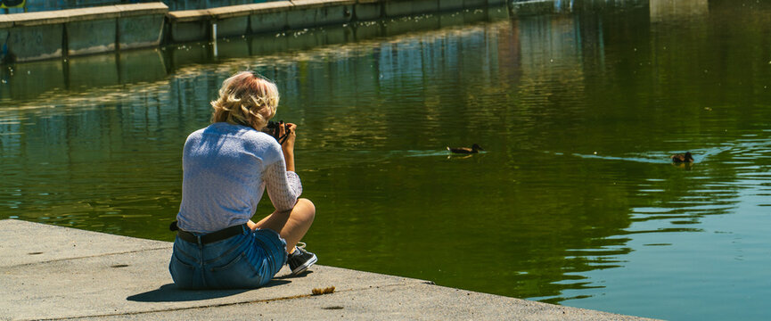 mujer sentada al lado de un lago haciendo fotos a los patos con su cámara réflex  , mujer joven con blusa blanca y pantalones cortos azules sujetando una cámara de fotos