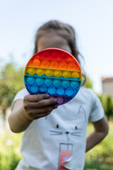 Naklejka premium The child is holding a rainbow pop it fidget toy instead of a head. Child plays with multicolored toy antistress pop it in the garden.