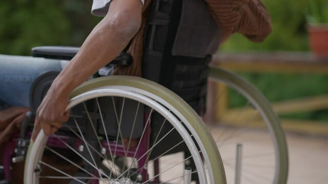 Camera moves up from wheelchair wheels to disabled African American man rolling outdoors on porch. Sad paraplegic adult guy riding manually operated device on summer spring day