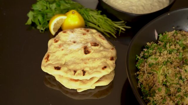 4K close up clip of woman's hand stacking homemade pitas into a pile on dark reflective table, other Greek foods can be seen in background. Indoors, kitchen, series.