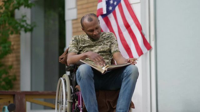 Portrait of handicapped African American veteran recalling memories looking through photo album sitting in wheelchair on porch with cigarette. Sad disabled man on backyard outdoors