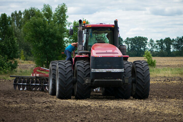 tractor driver near a large red tractor. A farmer on a tractor prepares land for a seeding tractor with a plow in an agricultural field. August, Ukraine