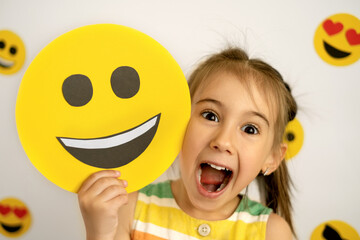 World emoji Day.  Smile Face. A little girl with a smiling cardboard smile face is laughing out loud with her mouth open and her eyes wide and bulging in surprise.
