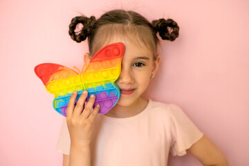 A beautiful cute little girl on a pink background covers one eye with a popit toy in the shape of a butterfly. Funny pigtails for a girl . Children's entertainment and educational toys. simple dimple
