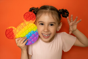An emotional portrait of a cute little girl on an orange background with a funny hairstyle made of pigtails and a popit toy  in her hands. Hand gestures. Pop it toy