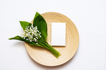 Stationary still life scene of Blank invitation or greeting card, Bamboo plate and lily of the valley flowers. Mockup, top view, flat lay