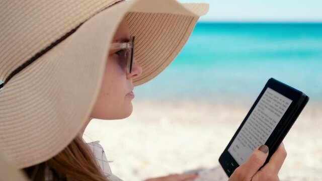 Close up of Woman in Hat Reading E-Book on Electronic Reader on Beach at Seaside in Summer Relaxing on Vacation in Hot Sunny Day. Digital Literature and Modern Leisure Concept. 4K Hand Held Orbit Shot