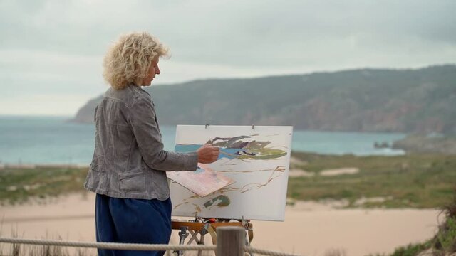 A middle-aged woman artist, by the sea, draws a beautiful sketch of a picture of a sea beach with mountains.