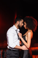Fototapeta premium Side view of man hugging african american girlfriend in dress on black background with red lighting