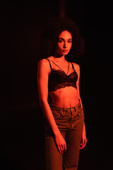 Young african american woman in jeans and bra looking at camera isolated on black with red lighting