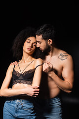 Shirtless man taking off lace bra from african american woman with closed eyes isolated on black