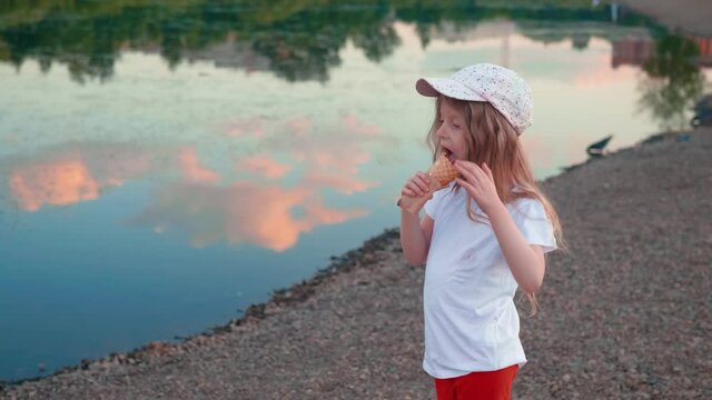 Side view little blonde gir with long hair licking ice cream next to river that reflects pink clouds. Cute child in white T-shirt eating waffle cone standing on the shingle bank.