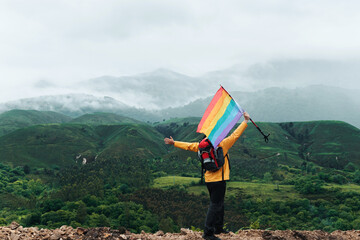 Obraz premium Hiker person on the mountain waving a rainbow gay pride flag. sport and inclusion. mountain activities- man with backpack doing trekking.