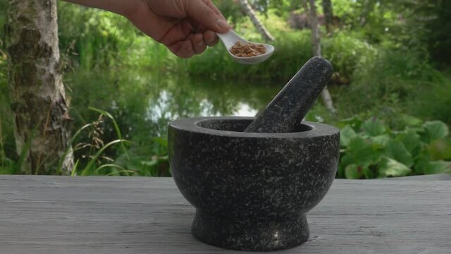 Hand is putting the spices into the grey stone mortar in slow motion outdoors