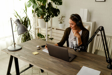 Woman with headphones sitting at table with laptop and microphone.