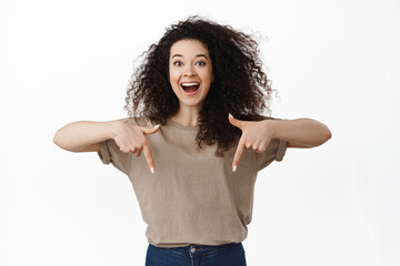 Excited beautiful woman showing announcement, pointing fingers down at advertisement and smiling amazed, happy to announce discounts in store, standing against white background © Cookie Studio