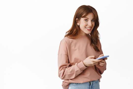 Image of smiling teenage girl with red hair, chatting on phone, messaging with smartphone and looking friendly at camera, standing over white background