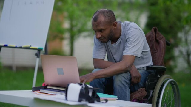 Portrait of concentrated handicapped African American man surfing Internet on laptop sitting in wheelchair on spring backyard. Professional disabled analyst with serious facial expression outdoors