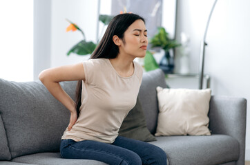 Upset frustrated asian young woman suffering from backache, unhappy japanese brunette sitting on a...