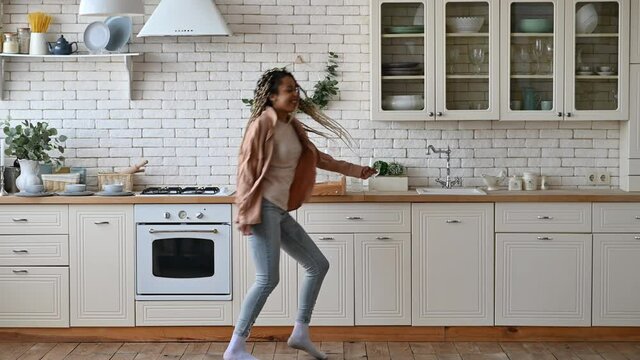 Cheerful happy young African American woman, in casual clothes, with dreadlocks, dancing at home in the kitchen to her favorite music, having fun at home alone