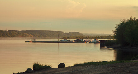 A deserted pier on the river on a sunny summer evening. The river is calm, the boats are moored, people in the distance are fishing.