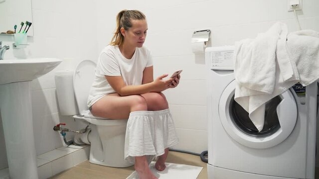 Happy beautiful blonde woman in pajamas smiles and holds smartphone in hands, sitting on the toilet. Chatting and gaming on the gadget. Lifestyle with phone. Using smartphone in everyday life
