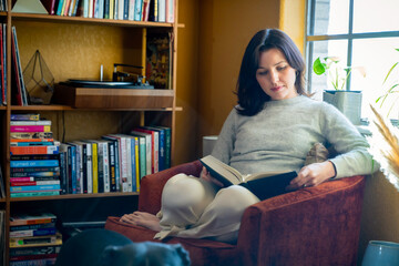 Woman sitting in orange chair by window and bookshelf reading a book