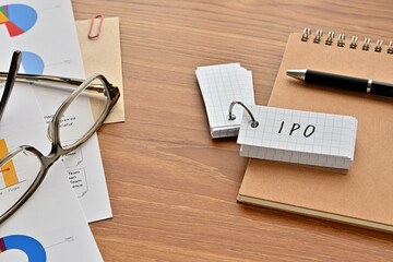 There is a word book with the word of IPO which is an abbreviation for initial public offering on the desk with papers of graphs(with dummy text), a pen and glasses.