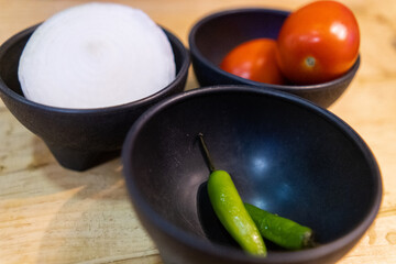 Tomatoes, onion, and chili peppers in black bowls above wooden surface
