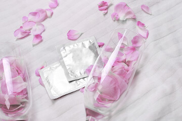 Condoms on white bed background. Glasses with flower petals, space for text. Romantic atmosphere with pink peony flowers. Date. Valentines day. Safe sex and reproductive health concept.