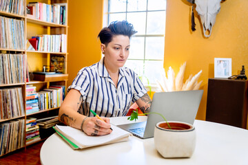 Masculine looking woman working from home on laptop, taking notes on notebook