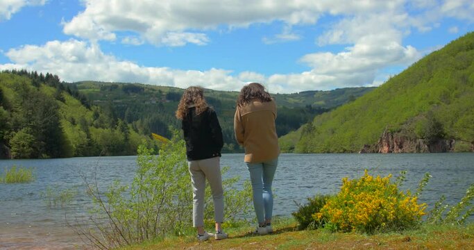 Young women with long hair, winter jacket turned away stand on lake waterfront and enjoy mountains nature view lake. Shot in 4k. Beautiful landscape of Loire, France