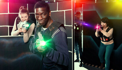 Cheerful African American man aiming laser gun at other players during lasertag game in dark labyrinth