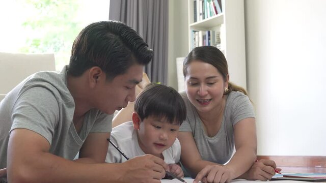 Asian family parents with little preschool son lying on the floor using color pencil drawing on paper book together in living room. Father and mother teaching child boy kid for homeschooling at home.