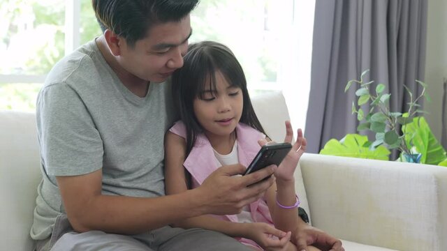 Asian family father and little daughter sit on sofa using smartphone playing game or watching cartoon together. Dad and child girl kid having fun weekend activity lifestyle with technology at home
