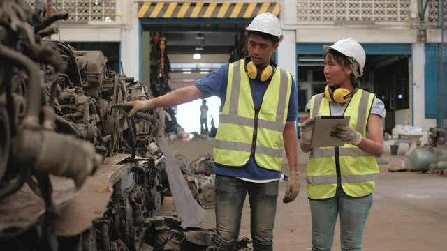 Asian male and female workers in uniforms and helmets, Employee stock check of car spare parts warehouse factory. Using a tablet to check old engines and machine orders for automotive industry.