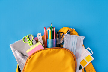 School supplies and medical mask with an antiseptic sticking out of backpack on blue background
