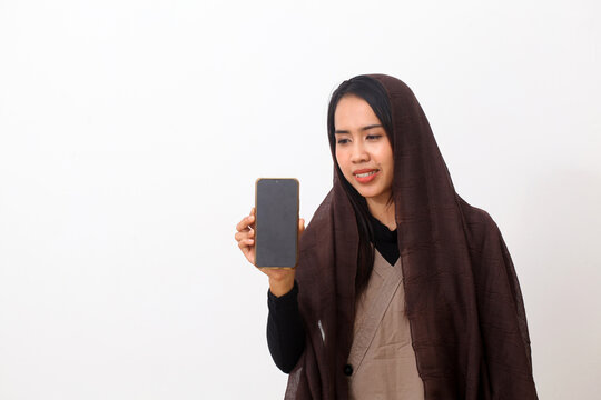 Portrait of happy asian muslim woman wearing a brown veil or hijab and white dress showing a blank smartphone screen. White background with copy space