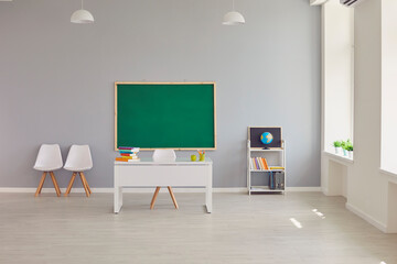 Interior of modern light empty spacious school classroom with clean green blackboard, teacher's desk, chairs, shelf, books and earth globe. Education, back to school concept