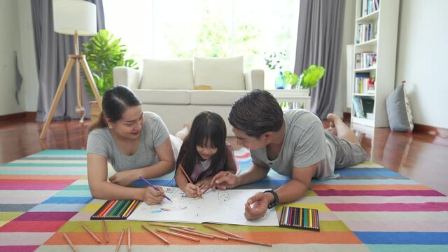 Asian family parents with little daughter lying on the floor using color pencil drawing on paper book together in living room. Father and mother teaching child girl kid for homeschooling at home.