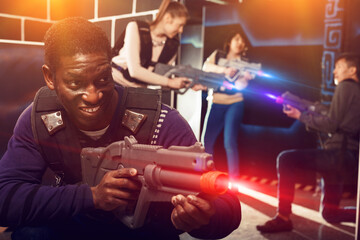 Obraz na płótnie Canvas Emotional African American man aiming laser pistol at other players during lasertag game in dark room