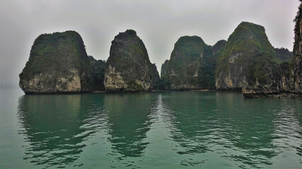 Naklejka premium Islands with bizarre outlines hide in the fog. Green vegetation on the steep slopes. Reflection on the water. Halong Bay. Vietnam